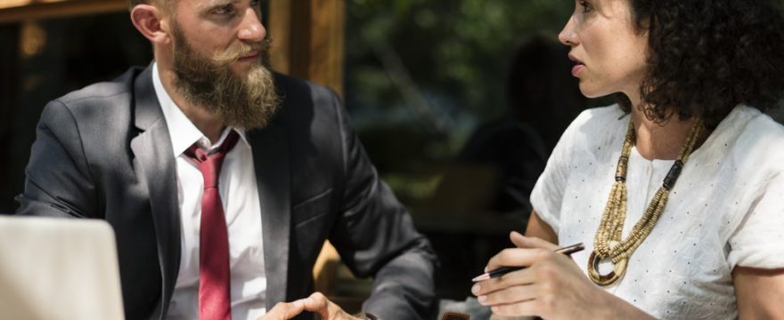 Man in a suit and woman in a white blouse are having a discussion outdoors at a table with a laptop and clipboard.