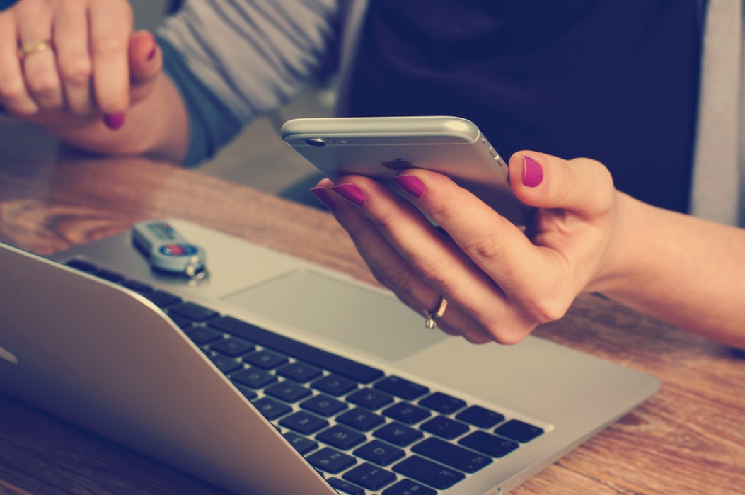 Person holding a smartphone, sitting at a desk with a laptop and a USB flash drive.