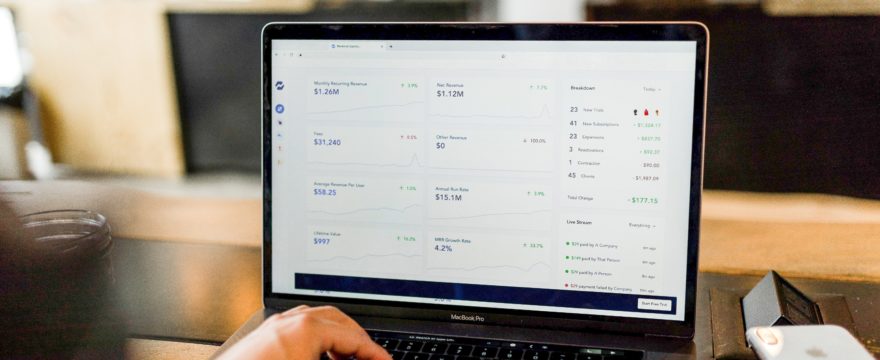 A person typing on a laptop displaying financial data and charts on the screen, sitting at a wooden table in a blurred background setting.