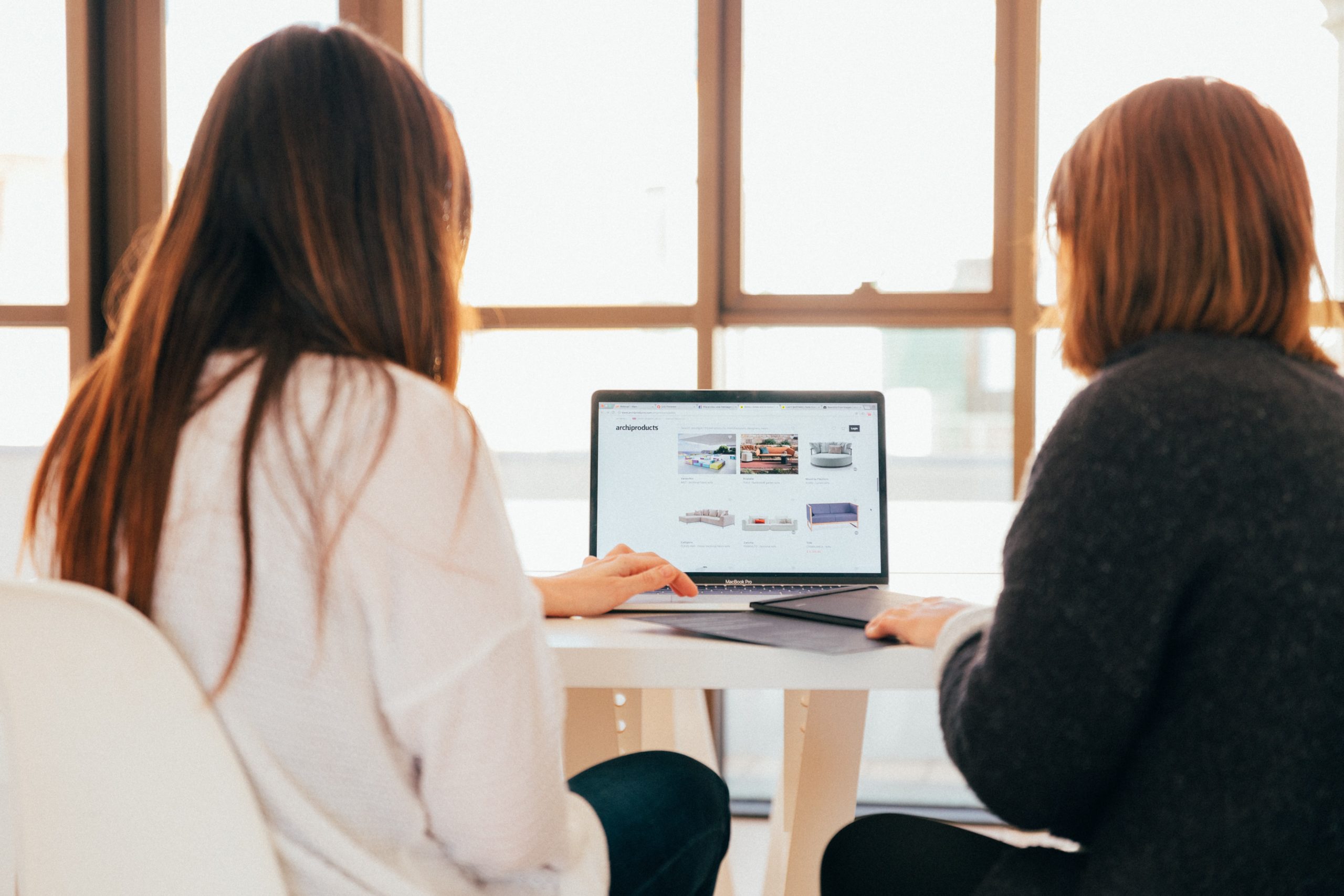 Two people sitting at a table, looking at a laptop screen displaying a travel website, with large windows in the background.