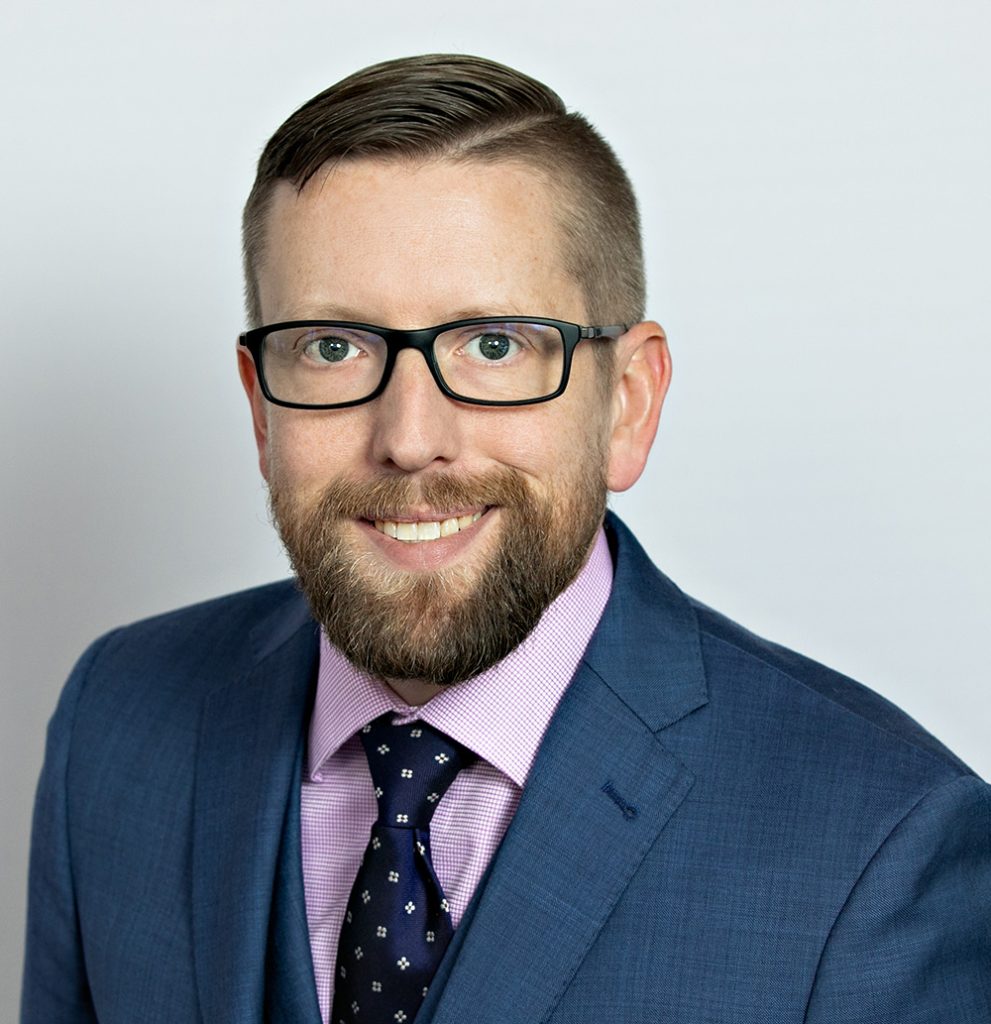 A bearded person wearing glasses, a blue suit, and a patterned tie, smiling against a plain background.