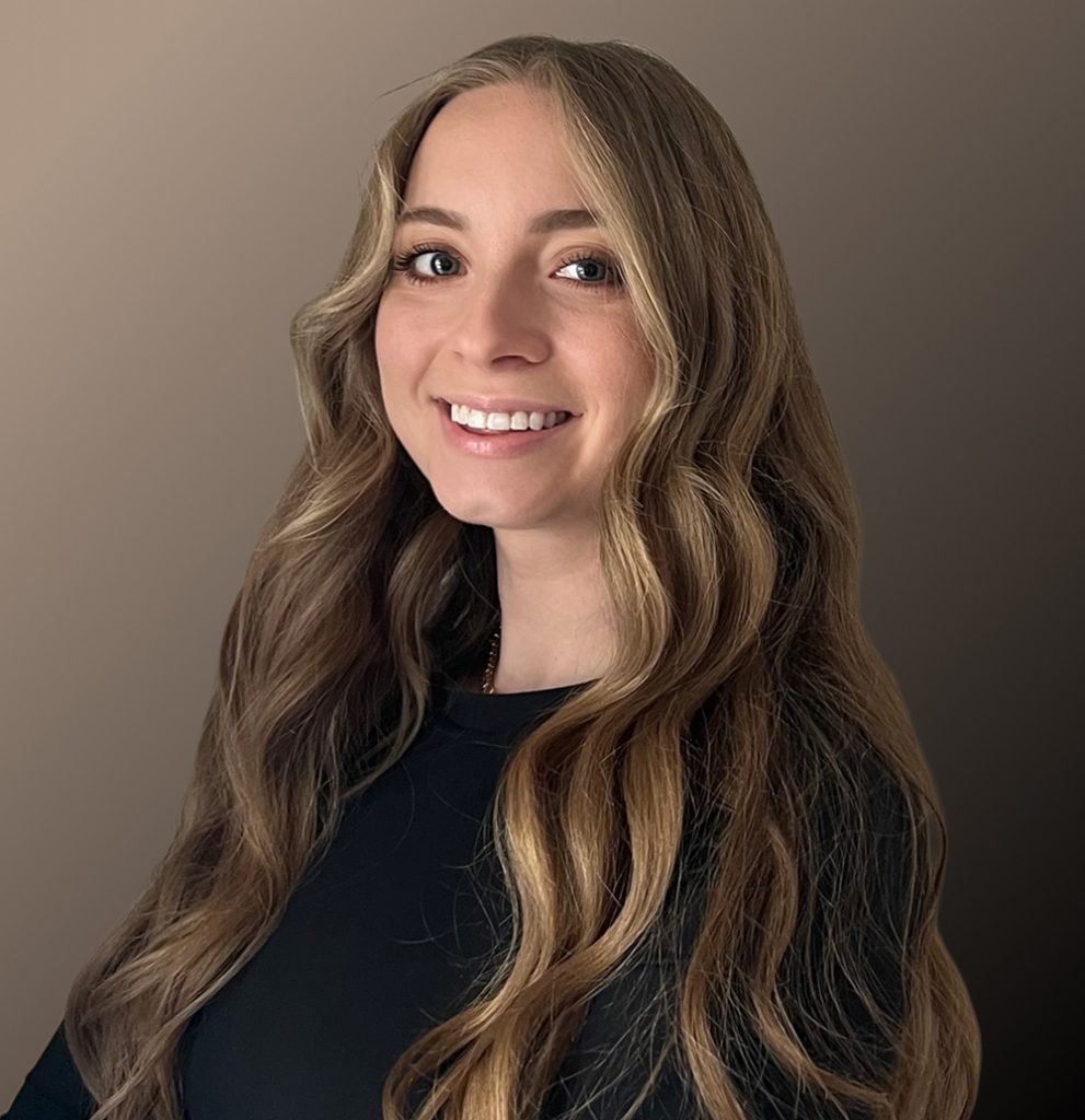 A person with long, wavy blonde hair smiles while wearing a black top, posed against a gradient brown background.