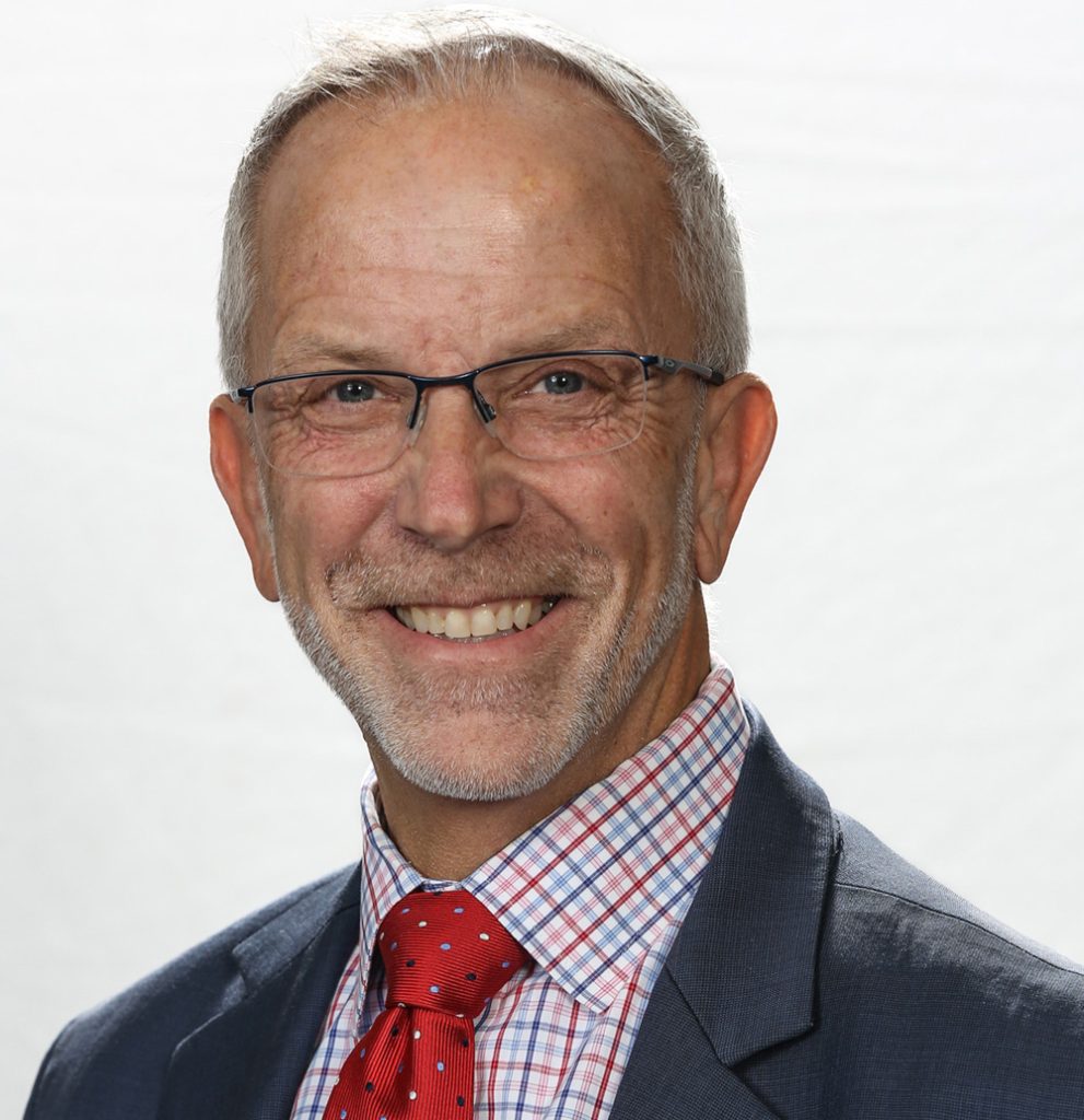 Smiling person with glasses, short hair, wearing a suit with a red tie and a checkered shirt, against a plain white background.