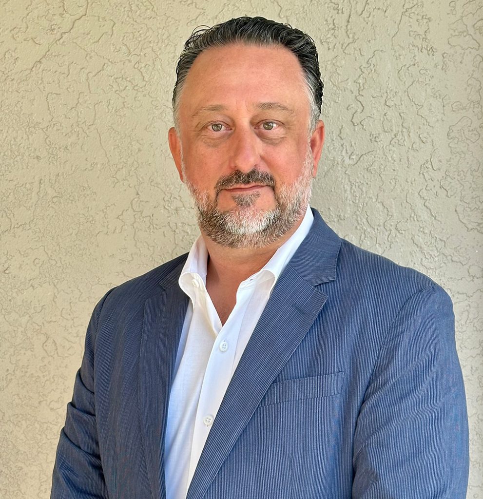 A person with a beard wearing a blue suit jacket over a white shirt stands against a textured wall.