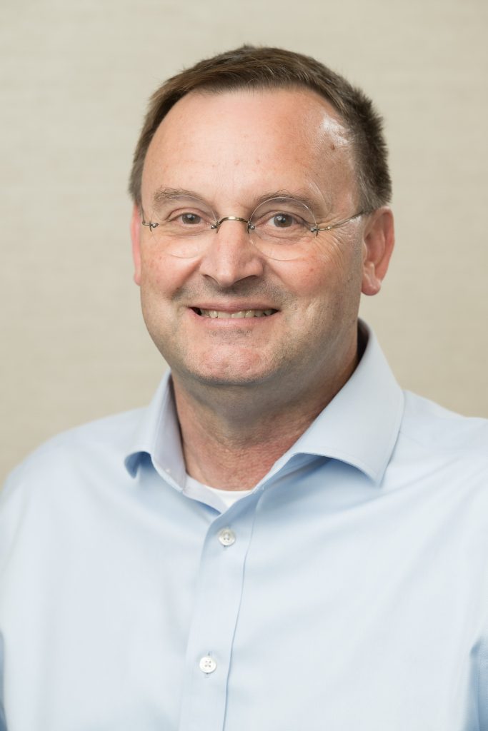 A middle-aged man with short brown hair and glasses, wearing a light blue collared shirt, smiles at the camera against a neutral background.