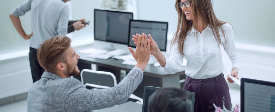 Two colleagues exchange a high five at a desk in a modern office, while another person works in the background.